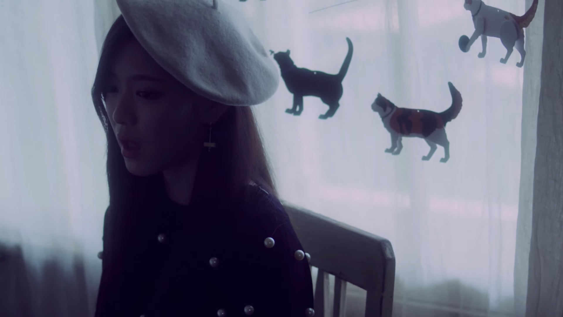 Hyunjin sitting in a chair, in front of a window with curtains decorated with cats.